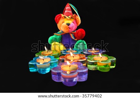 Happy Birthday teddy bear sits in front of flower shaped candles.  Candles are burning and in fun colors of blue, purple, green and orange.