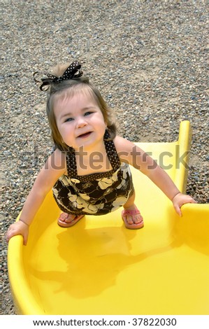 Baby crawls up the slide instead of sliding down the slide.  She is struggling upward but pauses to look up and smile.  Brown print sundress and sandals.