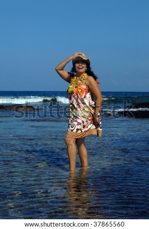 Beautiful woman stands in the waters of the Big Island of Hawaii.  She is wearing a swim suit cover-up, straw hat and lei.  She is tipping her hat to the bright sun and laughing.