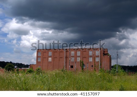 Old shut down factory is overshadowed by dark stormy clouds.  Overgrown with grass and weeds economic recovery seems bleak.