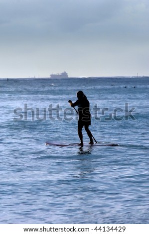 Older man paddle boards across Waikiki Bay while ship is leaving port.  Evening light silhouettes paddler.
