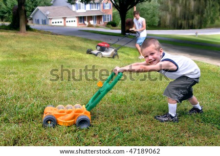 Small boy mows grass just like his dad.  He is grinning and pushing a toy lawn mower while dad mows with his.