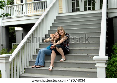 Mom and daughter sit on porch steps making faces and smiling.  Wooden steps have peeling grey paint.