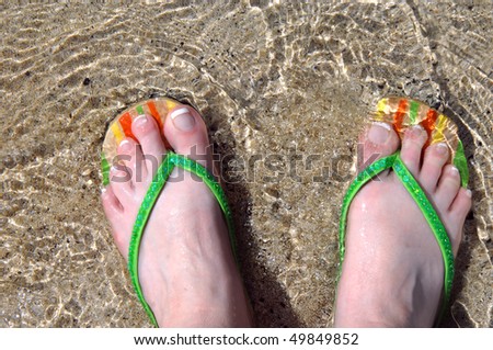 Woman\'s feet are covered with a gentle wave of water as she explores the beaches of the Big Island of Hawaii.  Flip flops are wooden with orange, yellow and green stripes.  Sequins line straps.