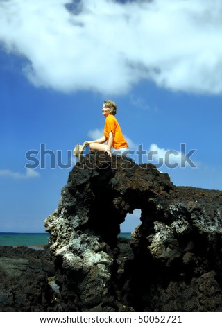Woman sits on top of a black lava rock arch on the Big Island of Hawaii.  She is holding her straw hat and feels on top of the world.