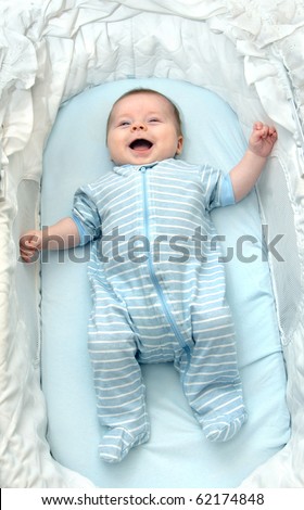 Infant smiles happily as he lies in an small baby bed.  He is wearing blue and white striped pajamas.