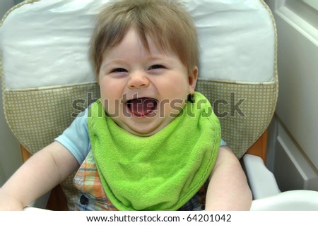 Infant smiles his widest smile ever as he laughs while sitting in his highchair in home kitchen.  He is wearing a bright green big.
