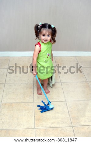 Adorable little girl pushes her mop back and forth cleaning the floors.  She is learning household chores from her mom.  She is wearing a green apron and holding a blue, play, mop.