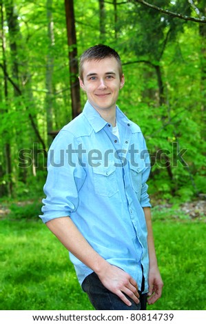 Young man stands surrounded by green forest.  He is wearing a light blue button front shirt and jeans.