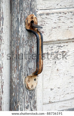 Rustic door handle, worn with much use, is mounted on a wooden, peeling painted, door.  Wood grain shows through and handle is rusty.