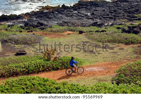 Biker takes the rugged trail along lava lined beach on Kauai, Hawaii.  Biker has on blue shirt and shorts.