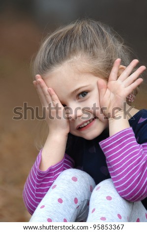 Small child is afraid to look as she peeps out from behind her hand.  She is sitting outdoors in a colorful purple striped shirt.