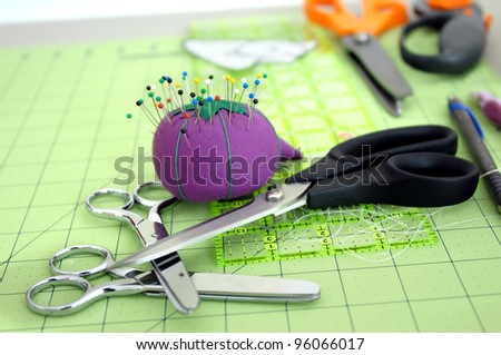Sewing tools of the trade lay on a green cutting matt.  Pins are stuck in purple pin cushion and three pair of different sized scissors lay in a stack on table top.
