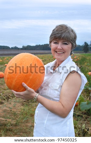 Older woman holds up a large pumpkin that she just picked from a pumpkin patch.  She is wearing a white shirt and smiling.  Field behind her has pumpkins ready for picking.