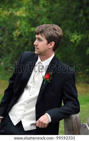 Groom, dressed in black tuxedo with with vest, shirt and tie, leans against a rustic wooden fence post.  He is solemn and thinking about his wedding day.