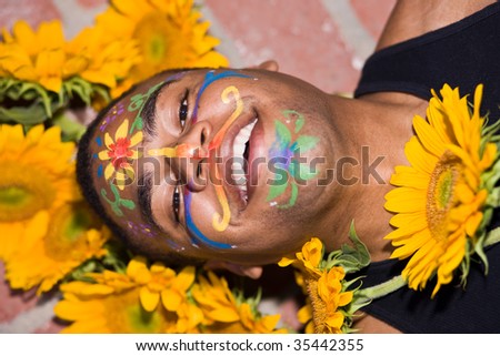 Young man with a painted face is laying on a patio surrounded by sunflowers.