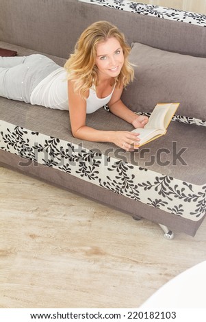 Pretty young woman enjoying reading a book at home lying on the sofa smiling in pleasure in casual clothing