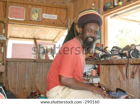 rasta man operating his shoe and leather repair shop on the beach in hillsborough, carriacou, grenada, west indies with photo of hailie selassi of ethiopipa on the wall.