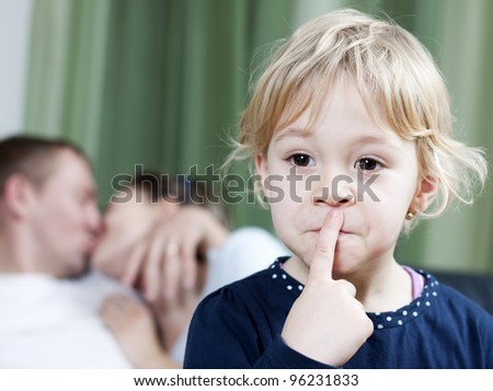 Blonde little girl making silence sign while mom and dad kissing in the background