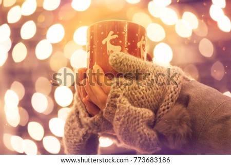 Young woman in winter gloves holding a mug of hot tea or wine outdoor, Christmas lights in the background
