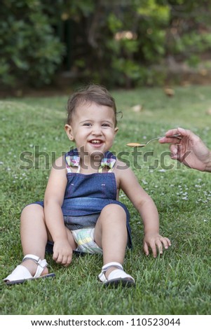 19-month baby smiling at camera WHILE hand of his mother feeds him, photograph outside a garden