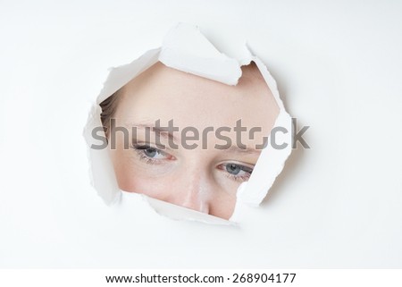 young woman peeking through hole torn in paper wall