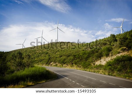 Aeolian generators of electricity on a hill. In first plane a road marks the diagonal of the frame. The sky, to the bottom, is blue intense with clouds. Horizontal frame