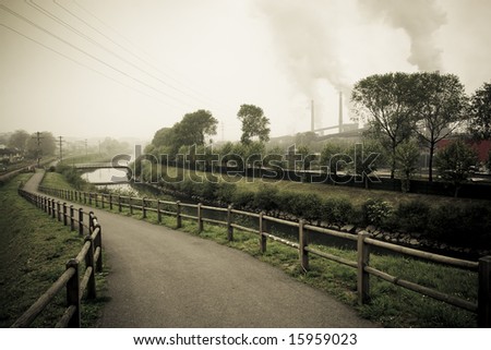 way reclaimed for pedestrian strolls near an industrial zone. It passes by the border of a river and is fence with wood. The bottom to the right the smoky chimneys of the industry are seen