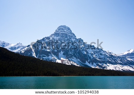 Rocky Mountain Peace -  a peaceful calm lake with a backdrop of coniferous forest and snow-capped mountain peaks under a blue sky.