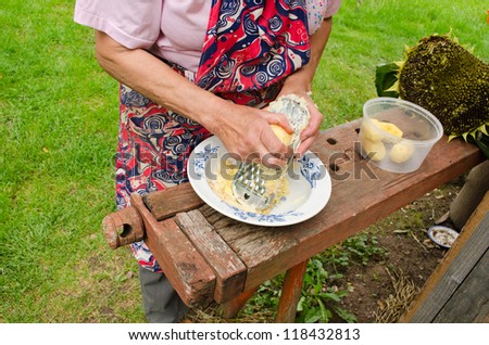 Old woman with apron hands grate peel potatoes with steel shredder tool on wooden outdoor table.
