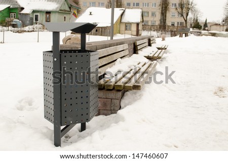 wooden park benches and waste bin covered with snow. winter town fragment and lithuania flag on house.