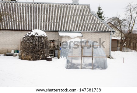 stack of firewood wood and polythene greenhouse conservatory covered with snow near white brick house in winter.