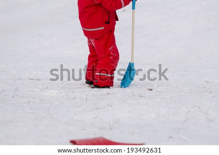 child stand with large snow shovel dressed in red waterproof clothing outdoor in winter time