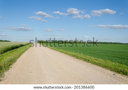 Rural gravel road between agriculture fields and wind mill windmill rorate generating alternative renewable electricity energy.