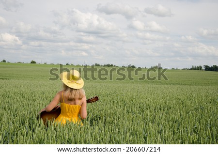 Country woman in yellow dress and hat on head play with guitar at wheat field.