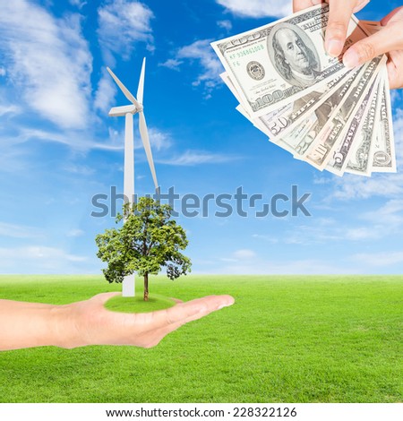 Carbon credits concept,hand holding wind turbine with tree and US Dollars banknote against green field and blue sky background
