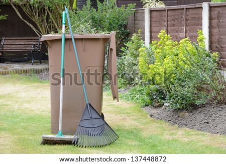 Stock image of garden waste in recycling container with lawn rake and sweeping brush in pleasant garden.