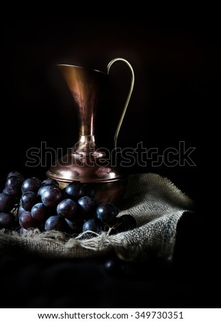 Creatively lit antique jug with black grapes against a dark rustic background with selective focus. The perfect cover image for your wine menu design.  Copy space.