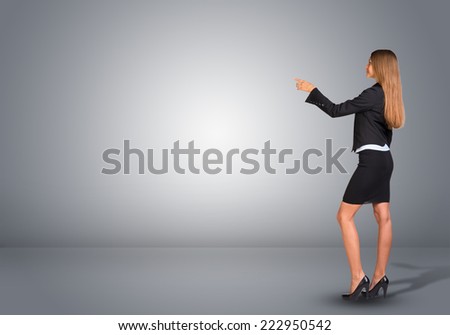Businesswoman in suit standing in an empty gray room and pointing finger at wall. Rear view