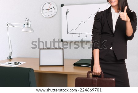 Businesswoman in suit holding briefcase and showing thumb-up. Office interior on background