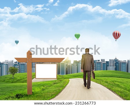 Businessman walks on road. Rear view. City skyline, grass field, wooden signboard and sky in background. Business concept
