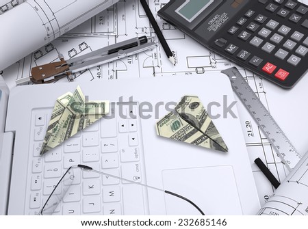 Paper airplanes of dollars lying on laptop keyboard. Architectural drawings and tools are close by. Concept of building business