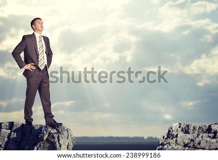 Businessman standing on the edge of rock gapm with arms akimbo. Sky and clouds as backdrop