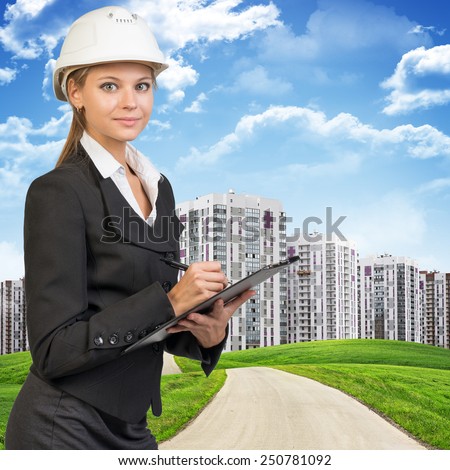 Businesswoman standing on the road among green hills, wearing hard hat, writing on clipboard, looking at camera. High-storey buildings, sky and clouds as backdrop