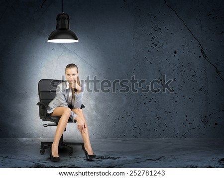 Businesswoman sitting on office chair in room in shell condition, lighted by lamp low above her head, looking at camera, smiling
