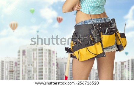 Woman in tool belt with different tools standing backwards, arms crossed. Cropped image. High-rise residential buildings and hot air balloons in background