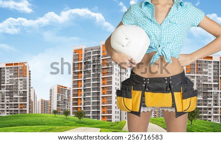 Woman in tool belt, shirt and jeans holding helmet under his arm. Cropped image. Green hills with road and buildings in background