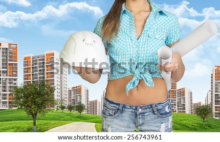 Woman in shirt and shorts holding white helmet and rolls of paper. Cropped image. Green hills with road and buildings in background