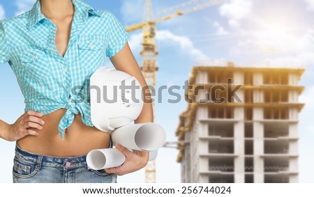 Woman in shirt and shorts holding white helmet and rolls of paper. Cropped image. Building under construction in background