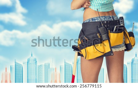 Woman in tool belt with different tools stands back, crossed arms. Cropped image. Wire-frame buildings as backdrop